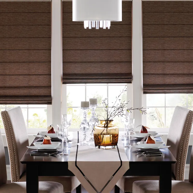 a dining room with a table set for a meal. There are four chairs around the table, and a centerpiece with a vase and branches. The windows have brown Roman shades.